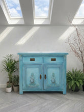 Load image into Gallery viewer, A hand-painted solid oak small sideboard or storage cabinet featuring a vibrant, multi-toned turquoise and sky blue blended finish. The piece has two top drawers with antique brass handles and two lower cupboard doors embellished with ornate, raised French-style scrolling appliques. The finish has a textured, aged patina, showcasing a blend of light and deep blues, styled in a bright room with indoor plants.
